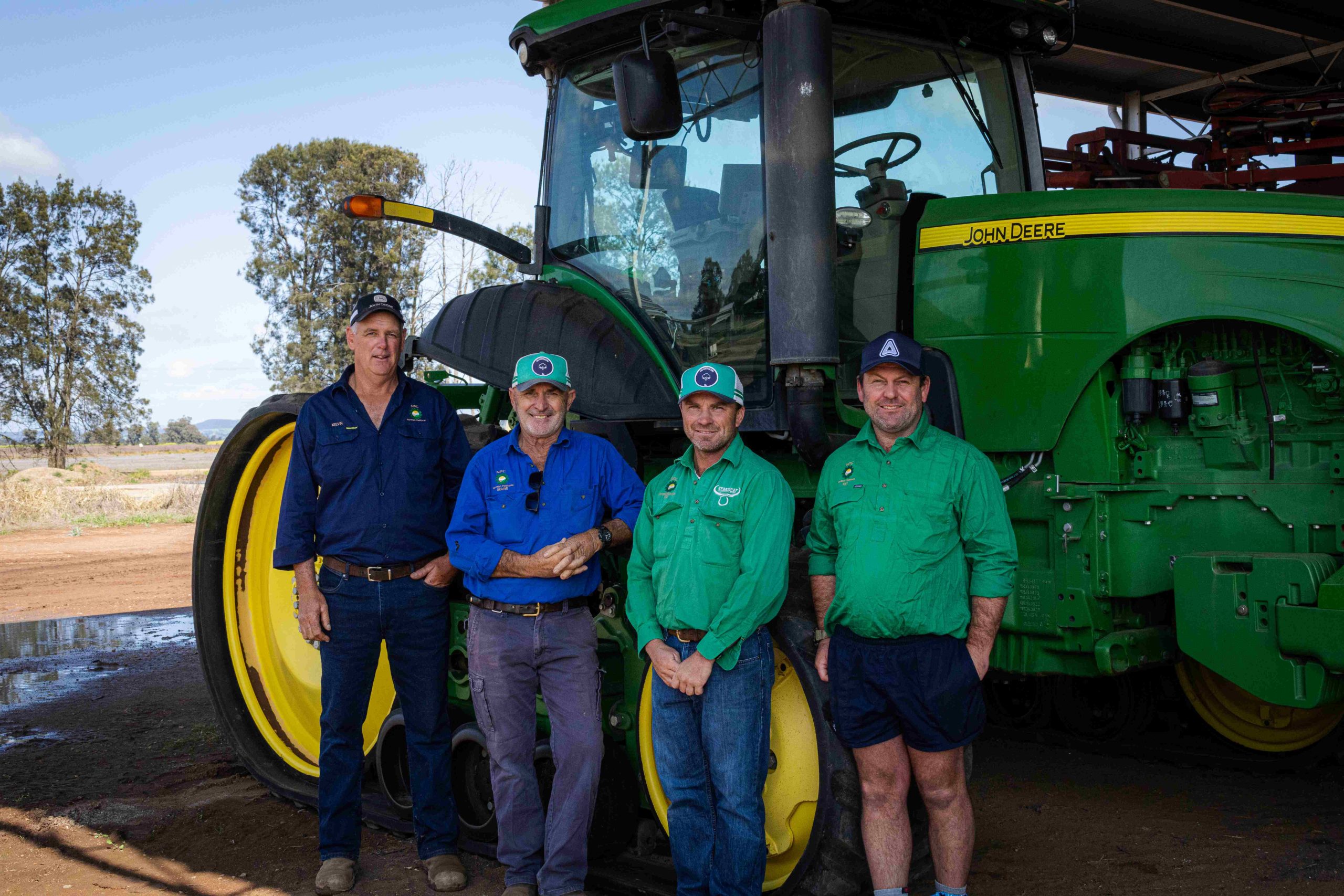 four farmers stand in front of green john Deere tractor in shed on sunny day